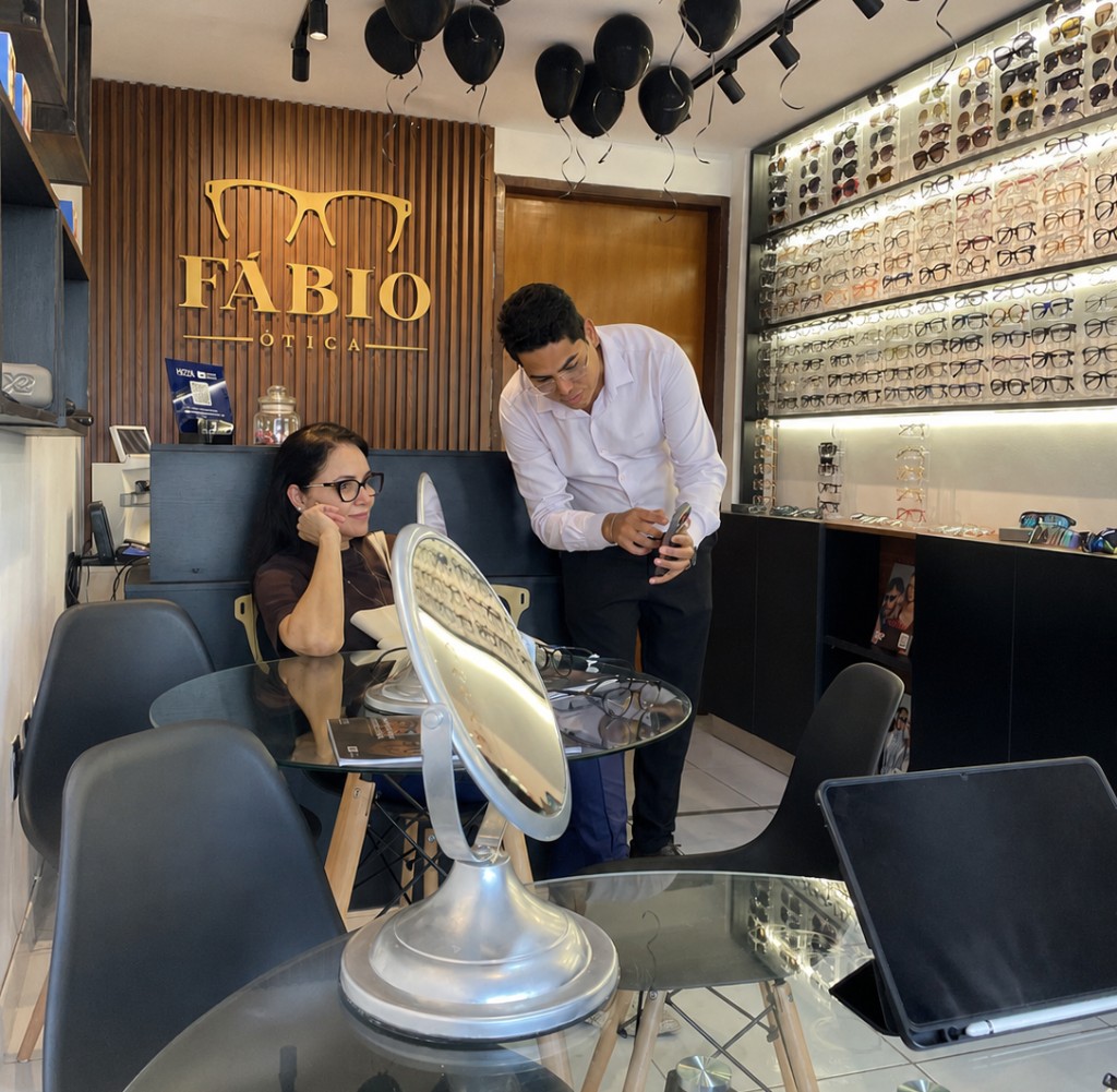 Atendimento na Fábio Ótica: cliente sentada à mesa de consulta e profissional ao lado, com expositores de armações ao fundo
